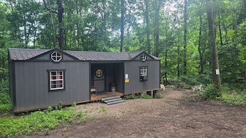 A small, dark-colored cabin with a single door and two windows, surrounded by a forest.