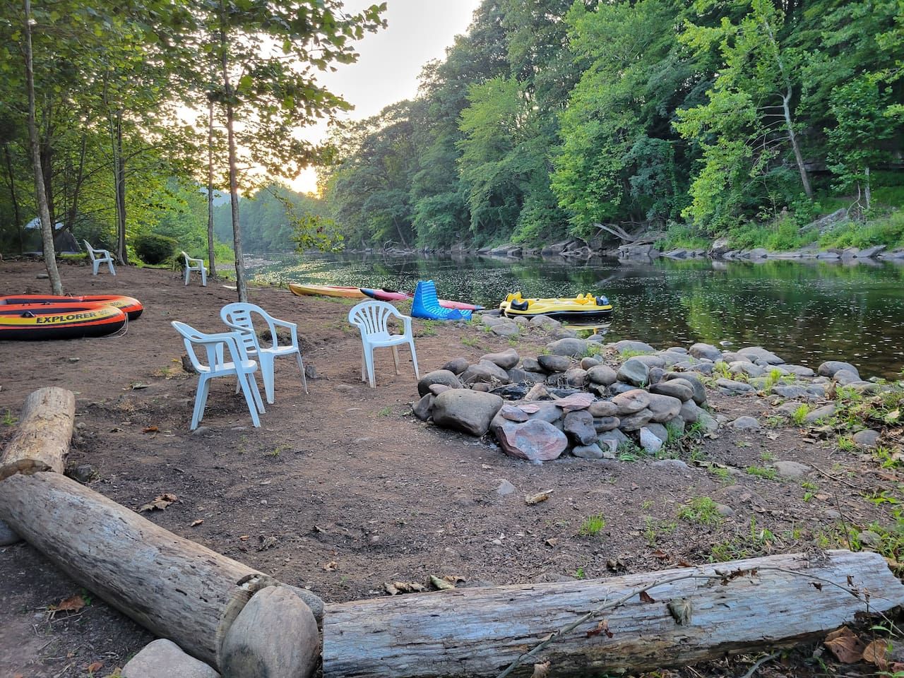 Un campamento con sillas, un montón de rocas y canoas junto a un río tranquilo rodeado de árboles.