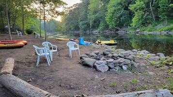 Un campamento con sillas, un montón de rocas y canoas junto a un río tranquilo rodeado de árboles.