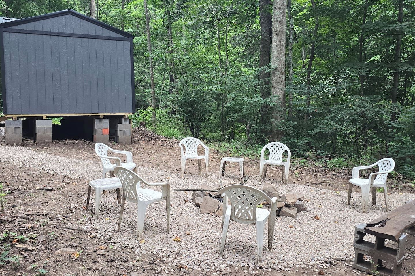 Un espacio de estar al aire libre rústico con sillas de plástico y una mesa de madera, rodeado de un entorno boscoso.