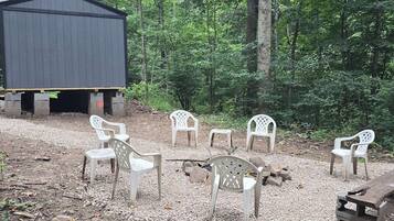 Un espacio de estar al aire libre rústico con sillas de plástico y una mesa de madera, rodeado de un entorno boscoso.