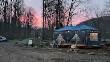 A blue tent with a table and chairs set up on a gravel area, surrounded by trees and a sunset in the background.