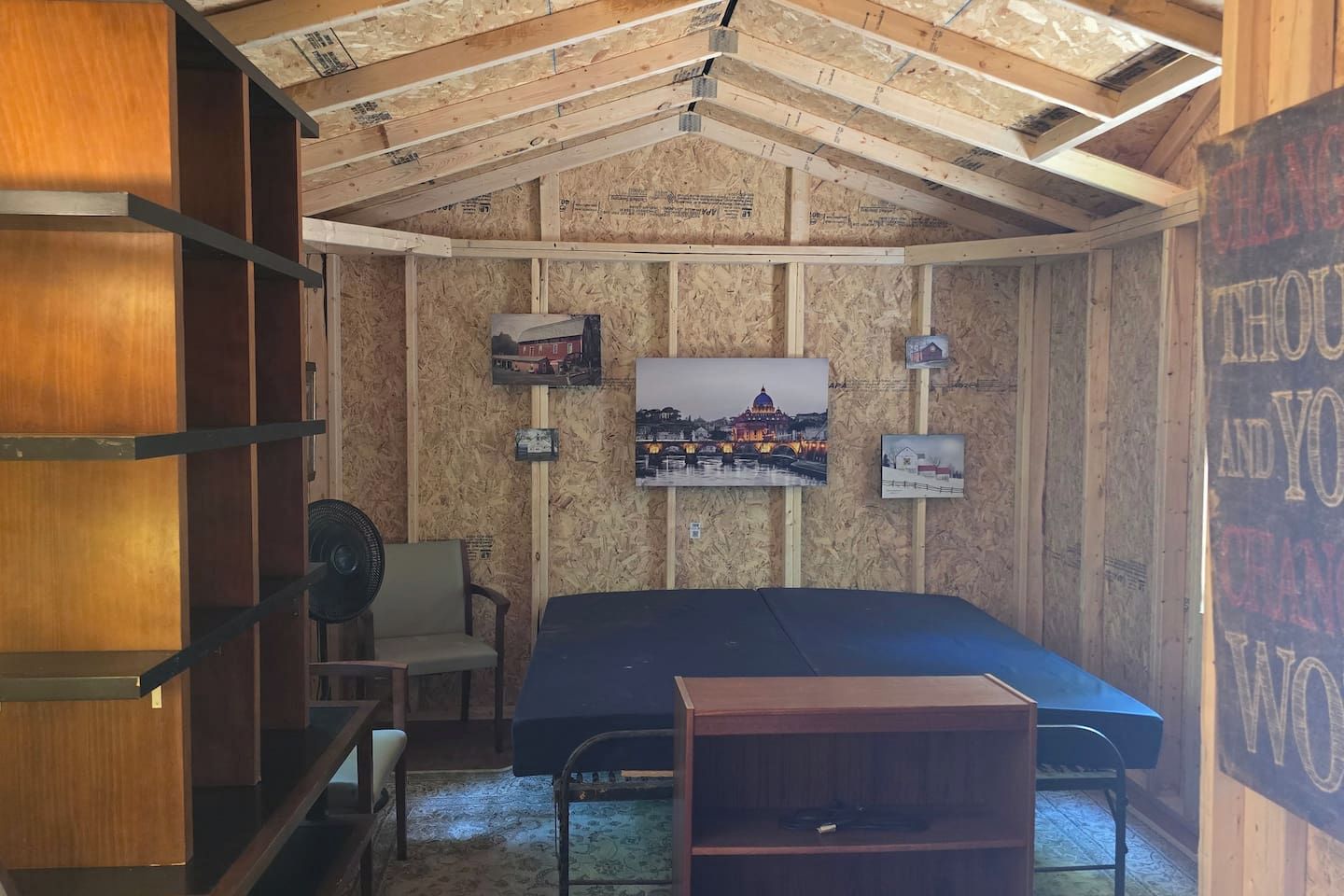 Una stanza con un letto, una sedia, un ventilatore e quadri appesi alle pareti.