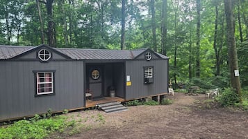 A small, dark wooden cabin with a porch, windows, and a door, surrounded by a forest.
