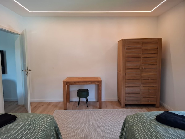 Una camera da letto con due letti, un armadio in legno, un piccolo tavolo con una sedia verde e una porta che dà su un'altra stanza.
