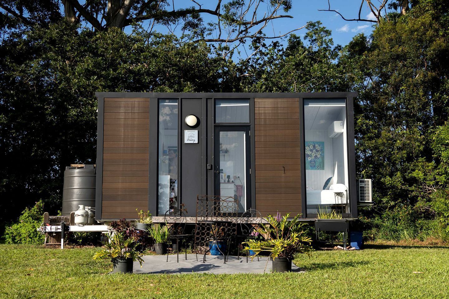 A modern, compact building with a large window, a door, and a small outdoor seating area with plants.