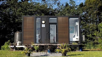 A modern, compact building with a large window, a door, and a small outdoor seating area with plants.