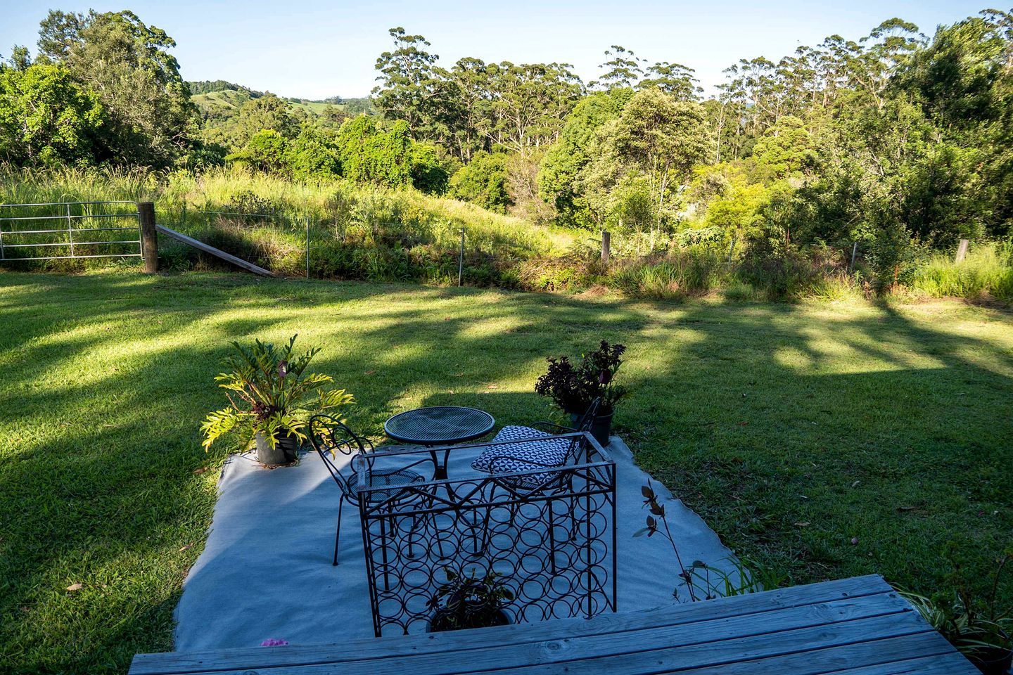 A grassy outdoor area with a metal table and chairs, surrounded by trees and a wooden fence.