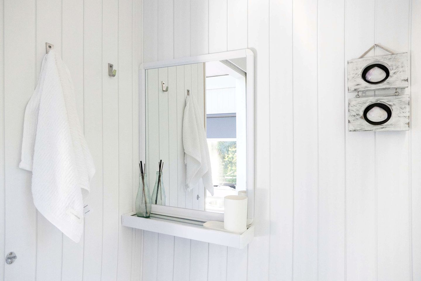 Une salle de bain avec un lambris en bois blanc, un miroir à cadre blanc, deux serviettes suspendues et une petite étagère sur laquelle se trouvent une bougie et deux bouteilles en verre.