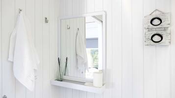 Une salle de bain avec un lambris en bois blanc, un miroir à cadre blanc, deux serviettes suspendues et une petite étagère sur laquelle se trouvent une bougie et deux bouteilles en verre.