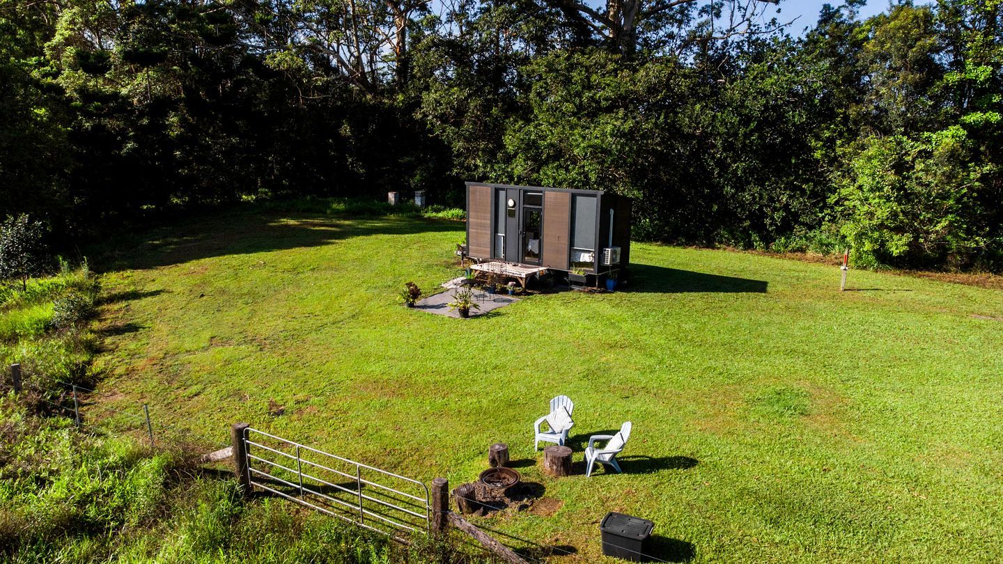 A small cabin with a door and windows, surrounded by a grassy area with a fence and two white chairs.