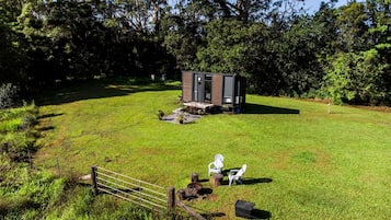 A small cabin with a door and windows, surrounded by a grassy area with a fence and two white chairs.