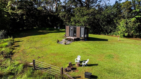A small cabin with a door and windows, surrounded by a grassy area with a fence and two white chairs.