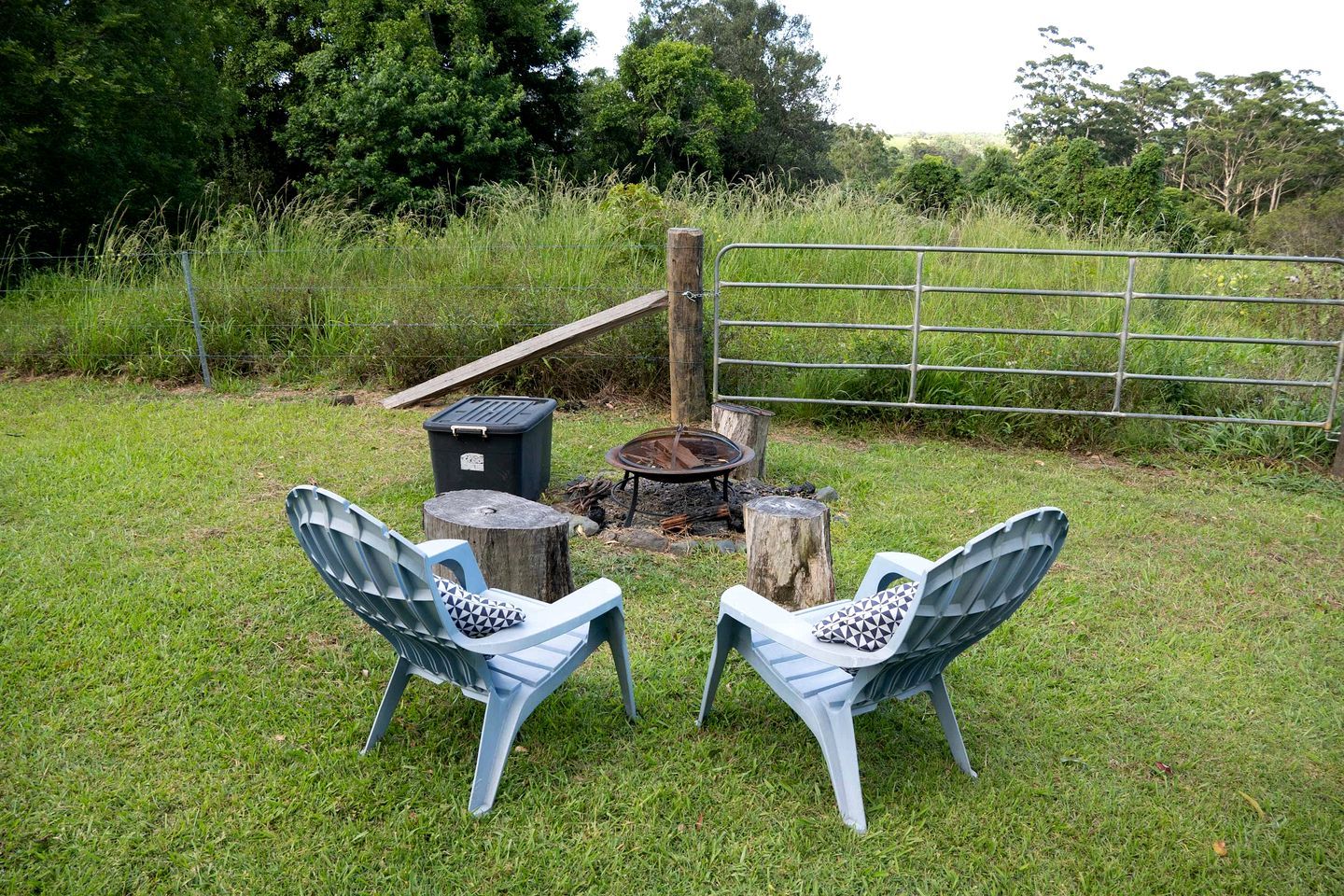An outdoor seating area with two chairs and a table, surrounded by a grassy field and a metal fence.