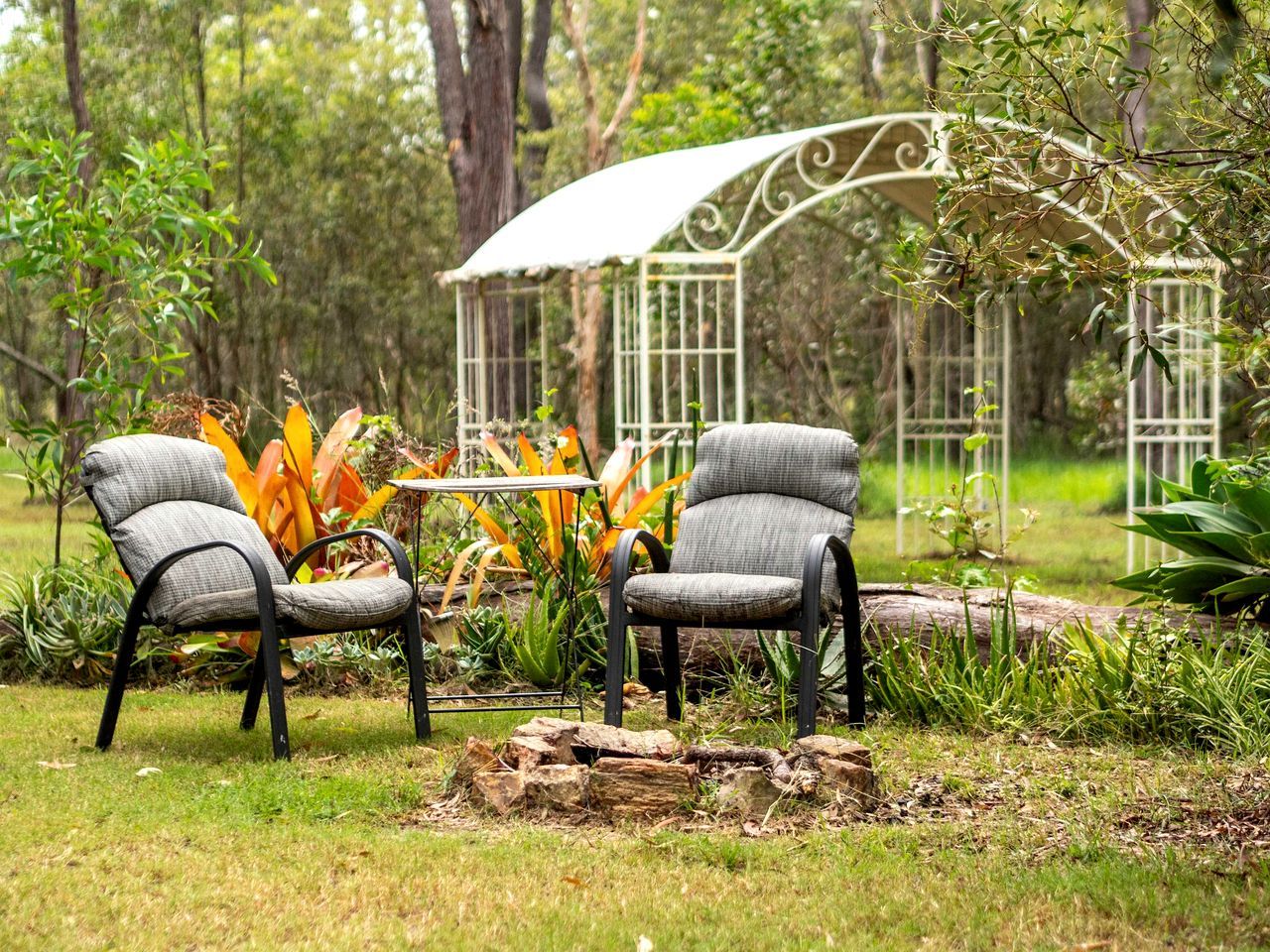 Two chairs with a table in a garden setting with a gazebo in the background.