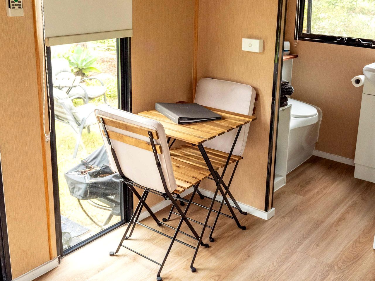 Une petite cuisine équipée avec une table et des chaises pliantes, une fenêtre donnant sur un espace verdoyant et des toilettes blanches.