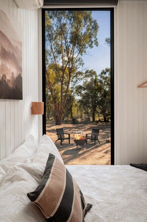 A bedroom with a bed, a view of an outdoor area with a fire pit and chairs, and a painting on the wall.