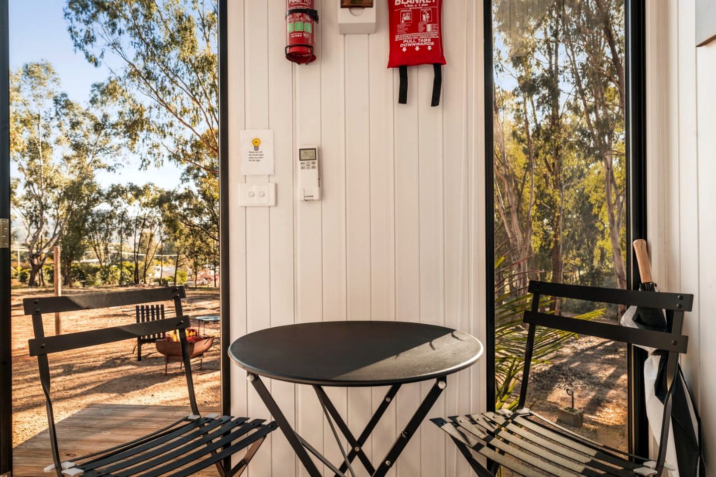 A room with a table and two chairs, a fire extinguisher, a blanket, and a window with a view of trees.