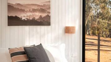 Un dormitorio con cama, almohadas, una colcha, aire acondicionado en la pared, un cuadro y vista a árboles por una ventana.