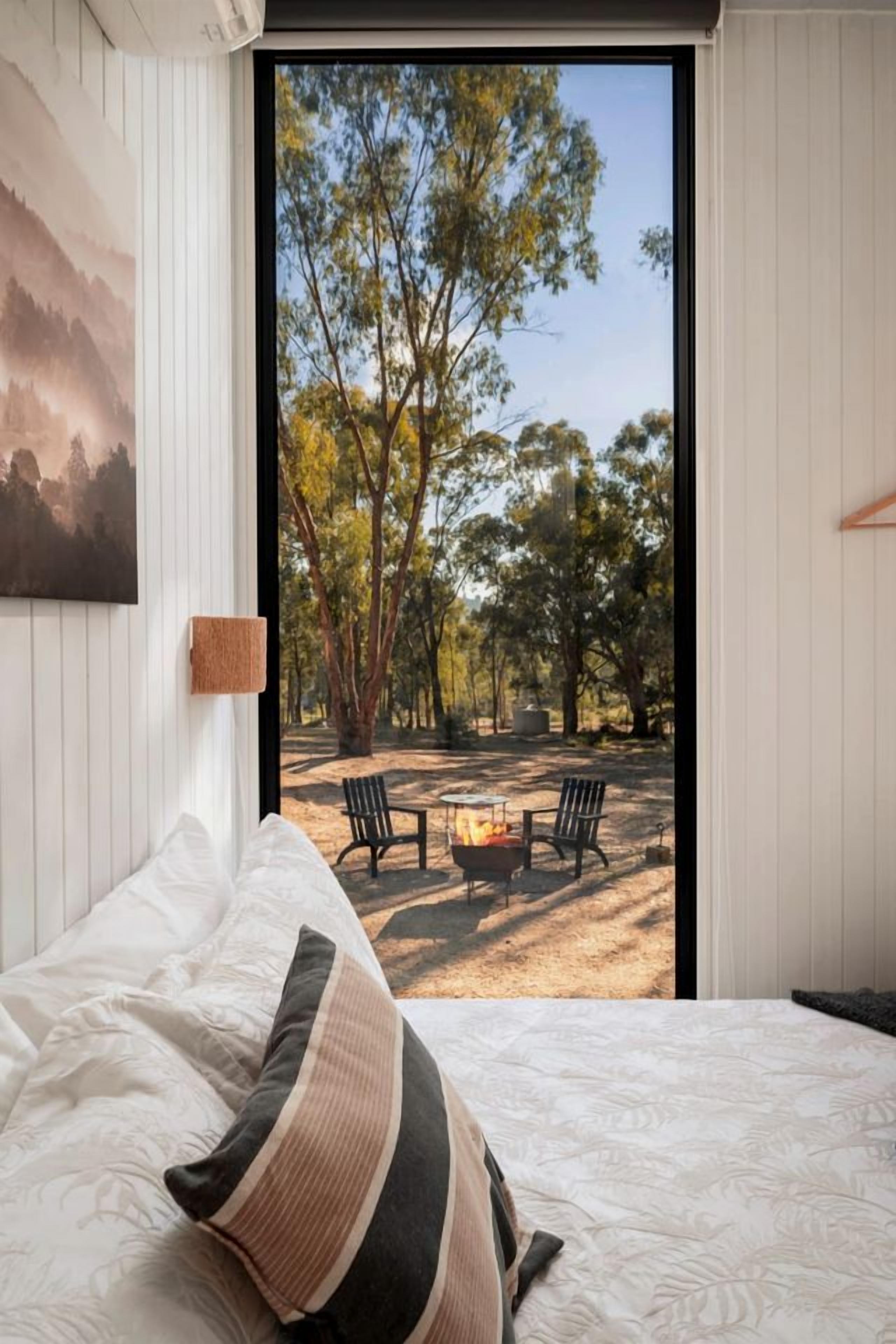 A bedroom with a bed, a view of an outdoor area with a fire pit and chairs, and a painting on the wall.