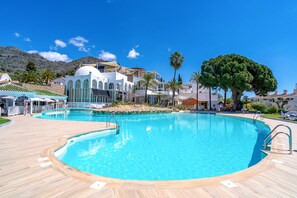 Ein großer Pool im Freien mit umlaufender Holzterrasse, ein weißes Gebäude mit Rundbogenfenstern und Palmen.
