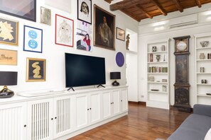 Ein Wohnzimmer mit einem Flachbildfernseher auf einer weißen Kommode, umgeben von verschiedenen gerahmten Kunstwerken, einer großen Uhr und einem Bücherregal.