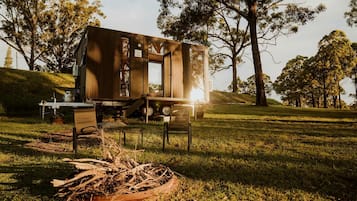 A rustic wooden structure with a window, a table, and chairs in a grassy outdoor setting.