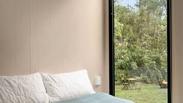 A bedroom with a bed, a window, and a view of an outdoor area with greenery and a fire pit.