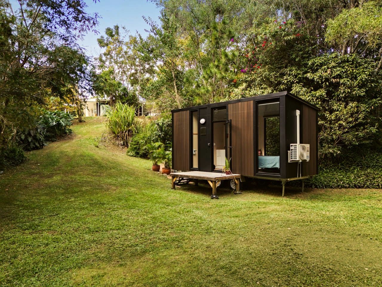 A small, dark-colored wooden structure with a door and windows, situated on a grassy area with trees and shrubs in the background.