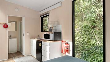 A compact room with a kitchenette, a bed, a window with a view of greenery, and a fire extinguisher.