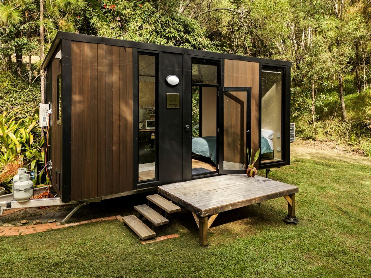 A compact wooden cabin with a deck, surrounded by greenery.
