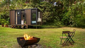 A modern, compact house with a fire pit in the foreground and a deck area with outdoor furniture.
