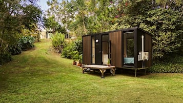 Une petite structure en bois de couleur sombre, dotée d’une porte et de fenêtres, située sur une zone herbeuse, avec des arbres et des arbustes en arrière-plan.
