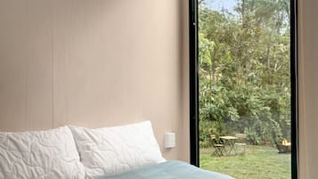 A bedroom with a bed, a window, and a view of an outdoor area with greenery and a fire pit.