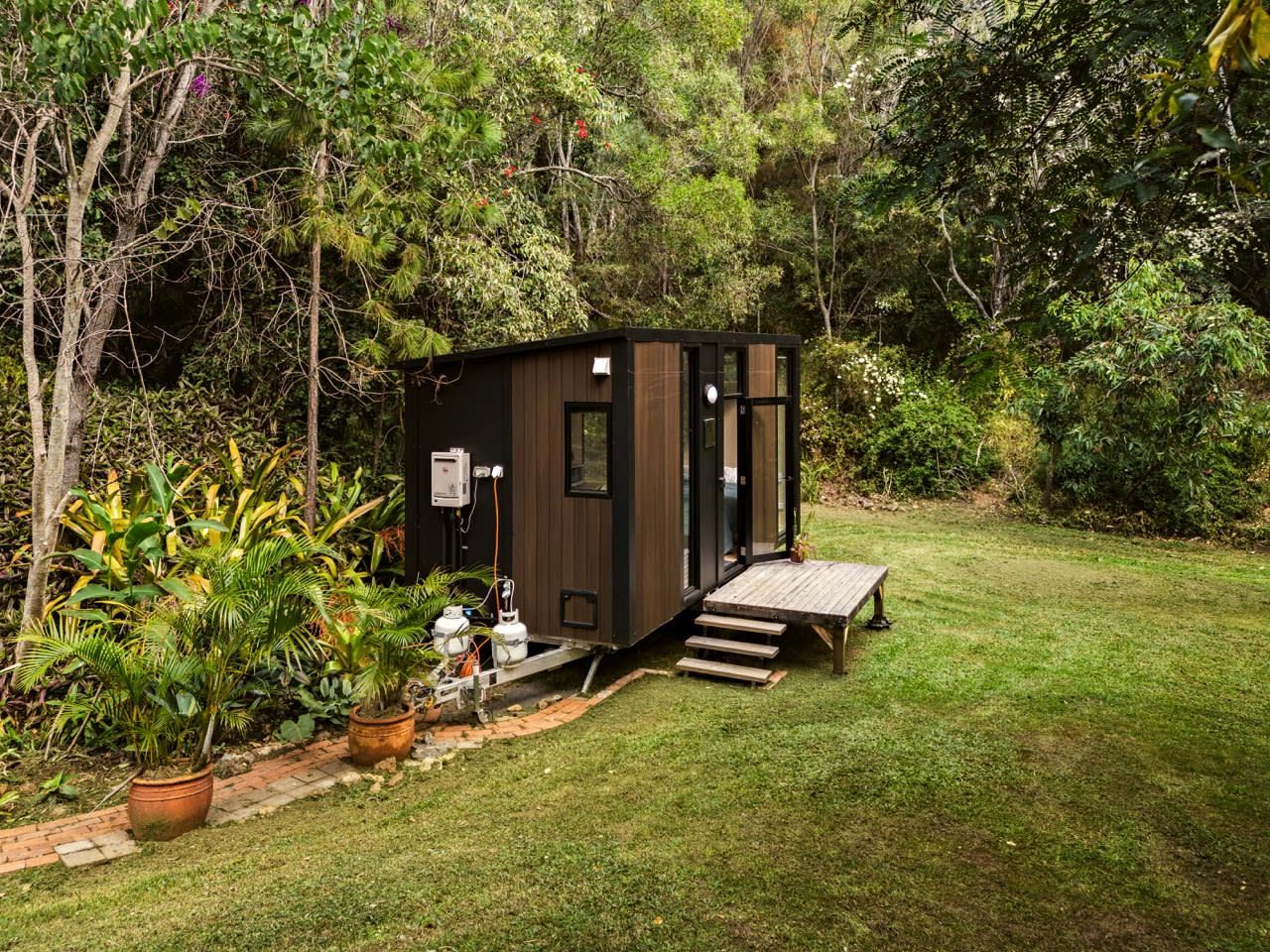 Een klein, donker vakantiehuisje met een houten terras en een trap er naartoe, omringd door weelderige begroeiing.