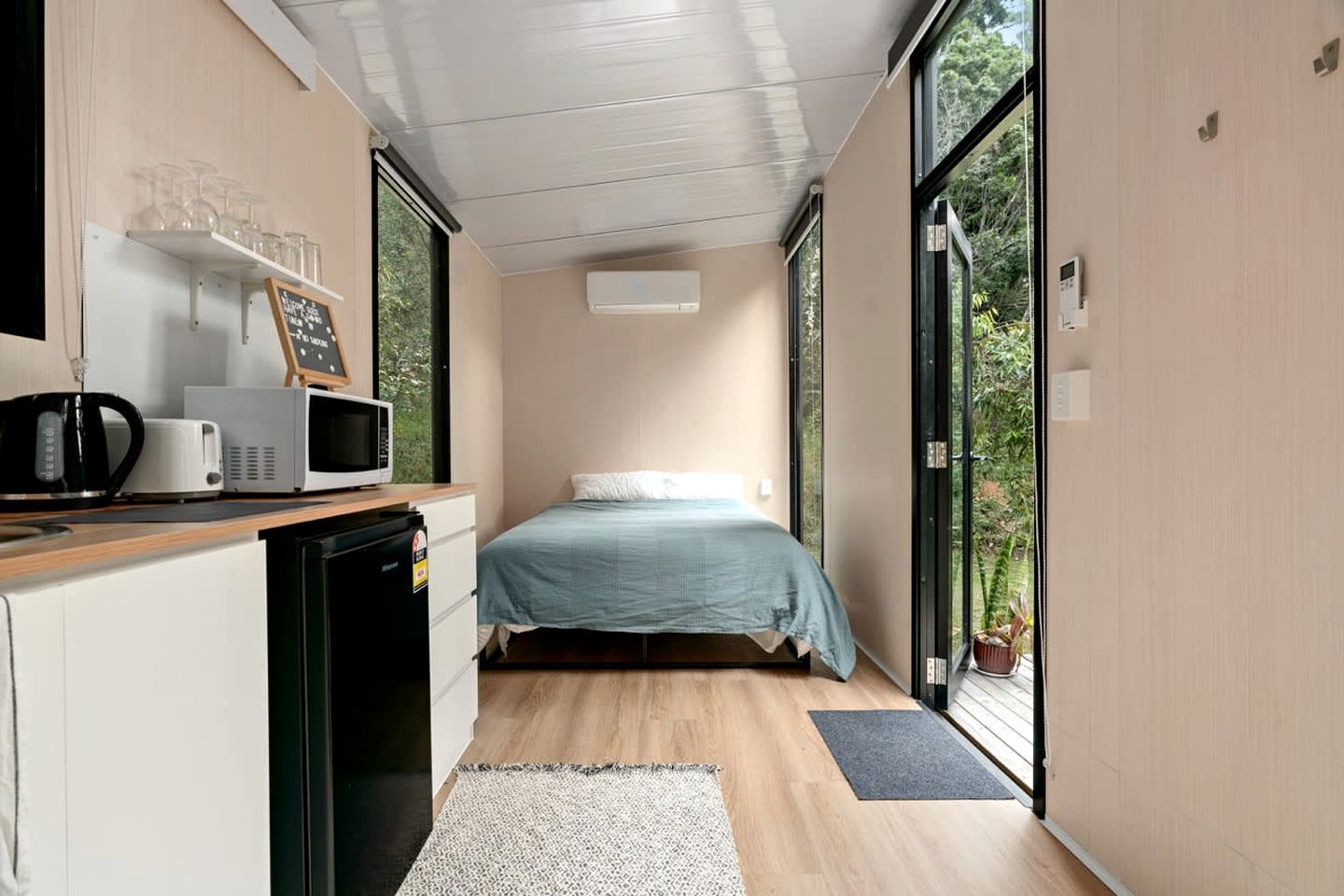 A bedroom with a bed, a desk with a microwave, and a view of greenery through the window.