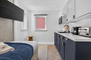Ein kleines Hotelzimmer mit einem Bett, einem runden Tischchen, einer Küchenzeile inklusive Mikrowelle, Toaster und Spüle sowie einem Fenster mit Blick auf Rosa.