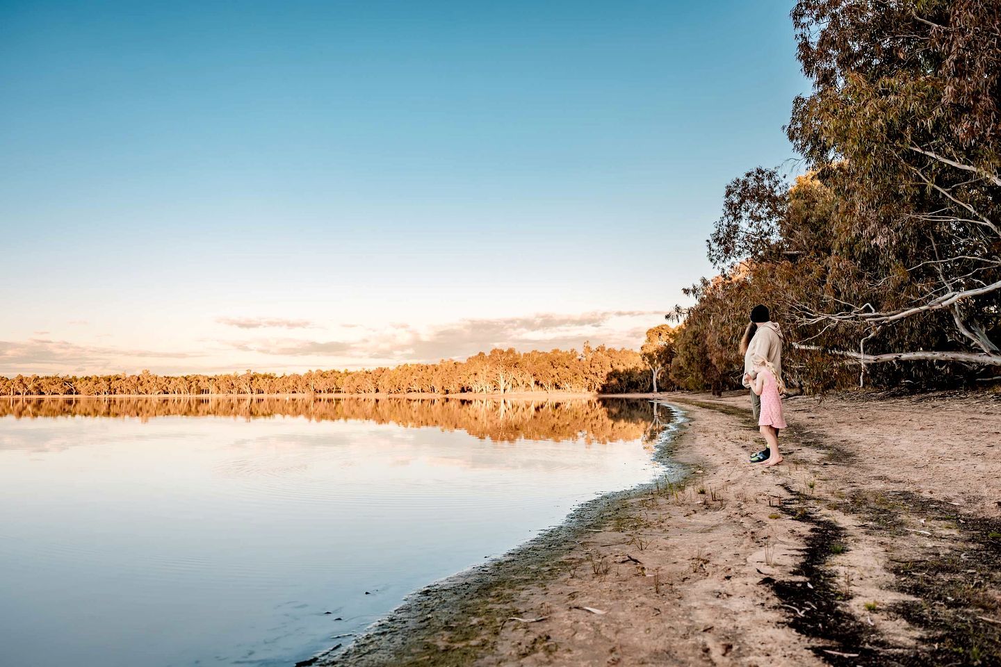 A person walking along a dirt path beside a calm lake with trees reflecting in the water.