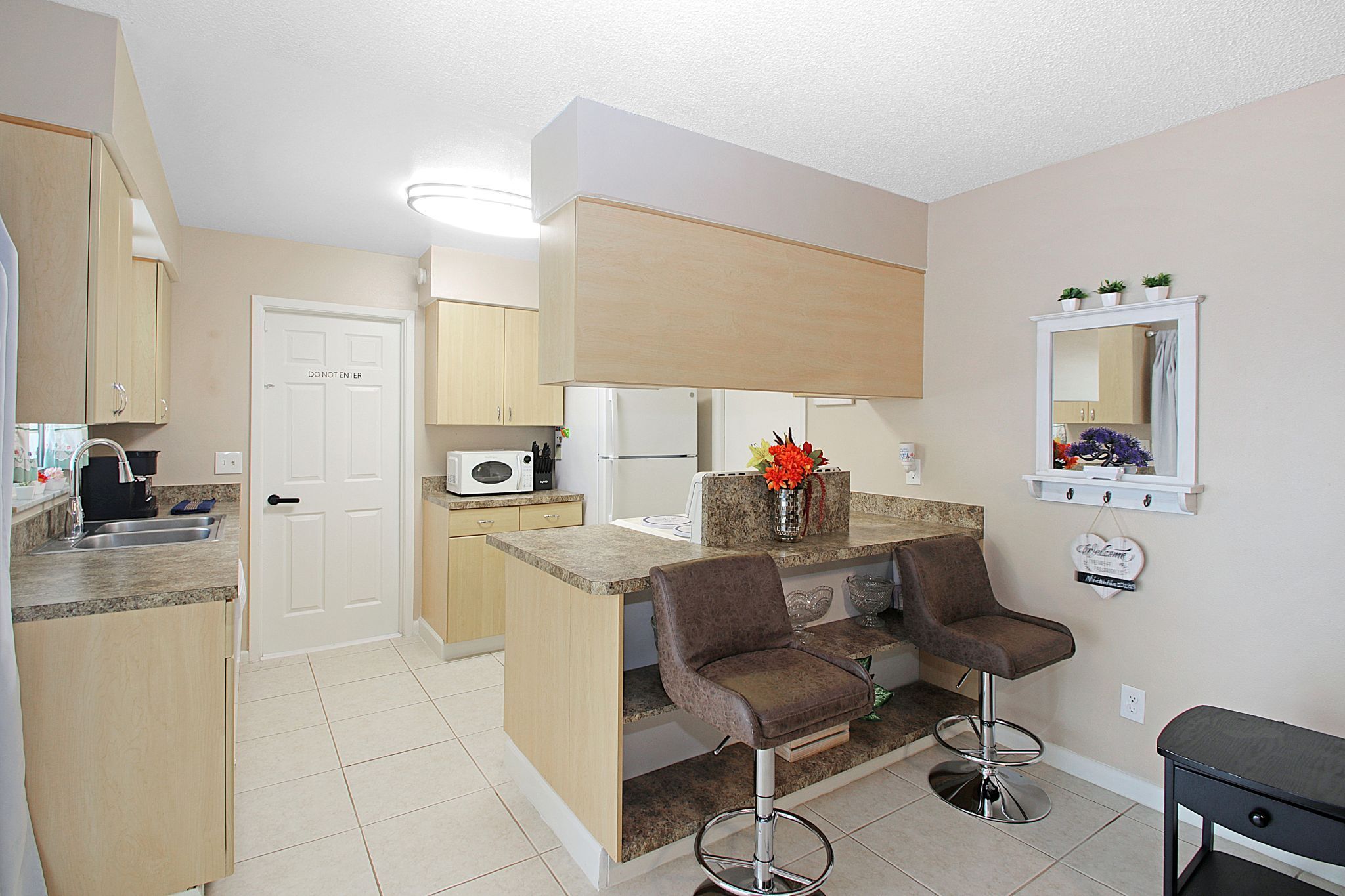 A compact kitchen with a microwave, a sink, and a door marked 'DO NOT ENTER.' There are two bar stools and a small table with a vase of flowers.