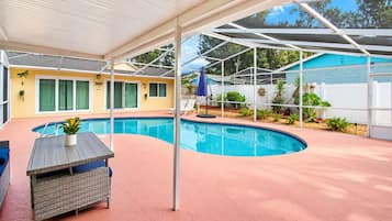 A covered outdoor swimming pool area with a lounge chair and a table, surrounded by a white fence and greenery.