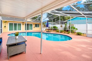 A covered outdoor swimming pool area with a lounge chair and a table, surrounded by a white fence and greenery.