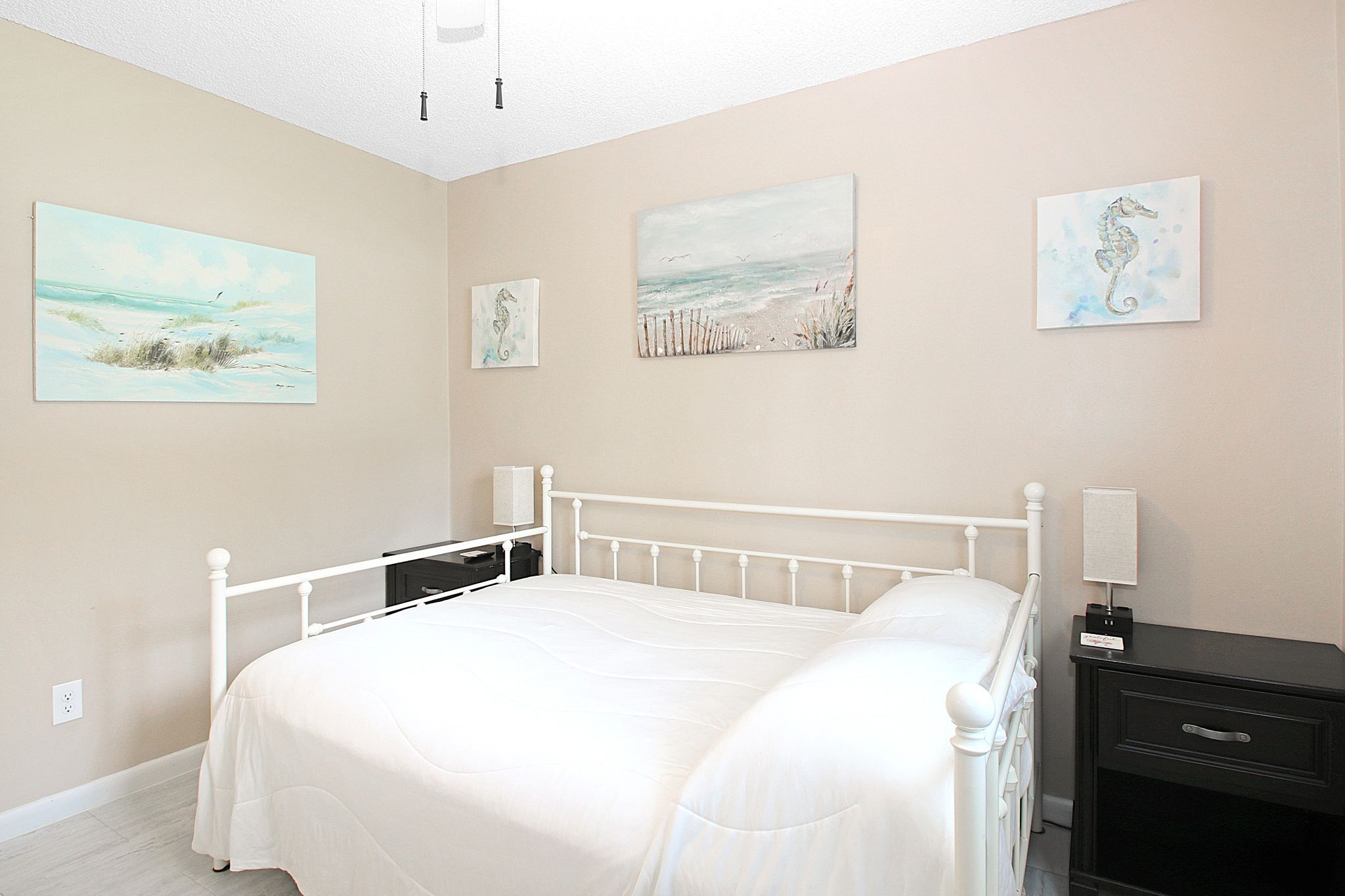 A bedroom with a white bed, a nightstand, and a dresser. There are three paintings on the wall.