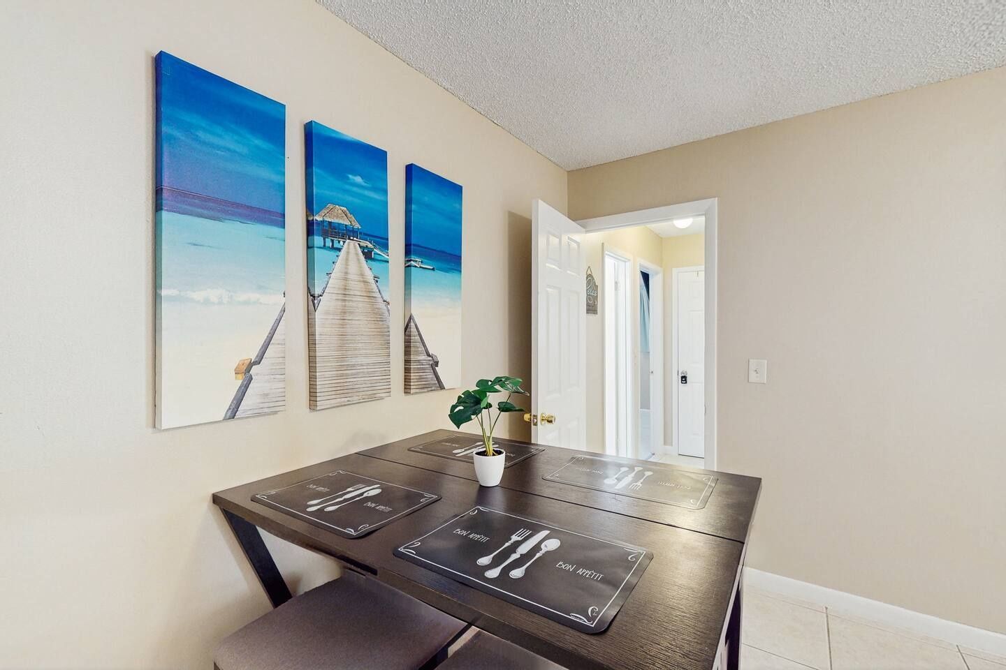 A dining area with a table set for four, a potted plant, and three wall-mounted paintings depicting a coastal scene.
