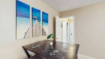A dining area with a table set for four, a potted plant, and three wall-mounted paintings depicting a coastal scene.