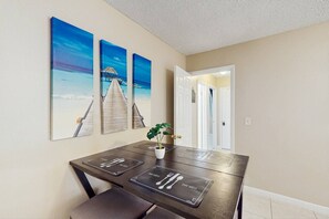 Un coin repas avec une table dressée pour quatre personnes, une plante en pot et trois tableaux accrochés au mur représentant un paysage côtier.