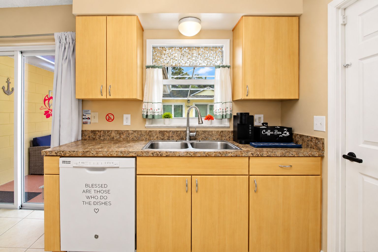 Una cocina compacta con lavavajillas, cafetera y ventana con cortina decorativa.