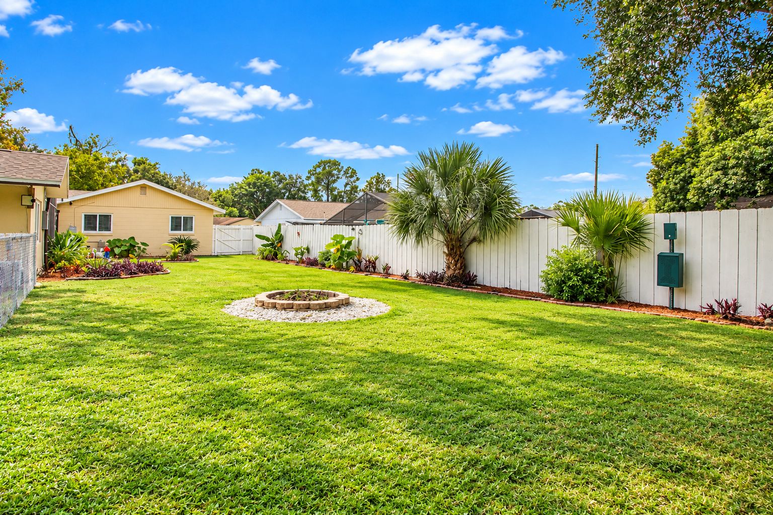 A well-maintained backyard with a circular fire pit, a white fence, and a variety of plants.