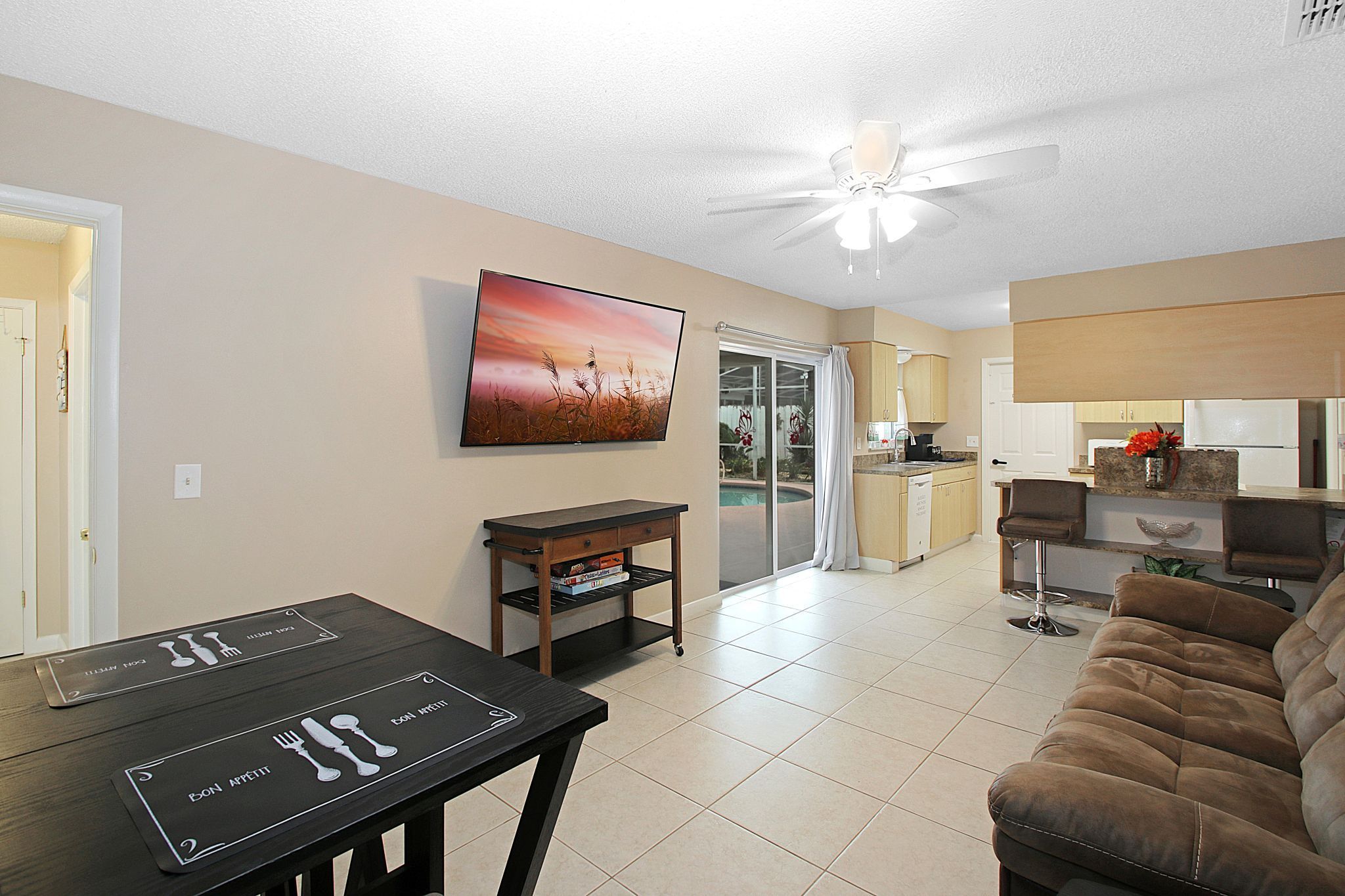 A modern living room with a dining table, a sofa, a ceiling fan, and a kitchen area with a sliding glass door leading to a patio.