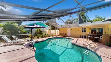 A swimming pool with a glass enclosure, lounge chairs, and a patio area with a table and chairs.