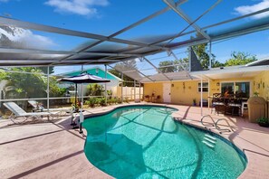 Une piscine entourée de parois en verre, des chaises longues et un espace patio avec une table et des chaises.